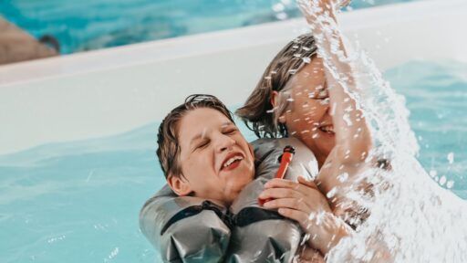 Eine ältere Frau planscht mit ihrem Enkel in einem Schwimmbad, der Wasser spritzt, beide lachen.