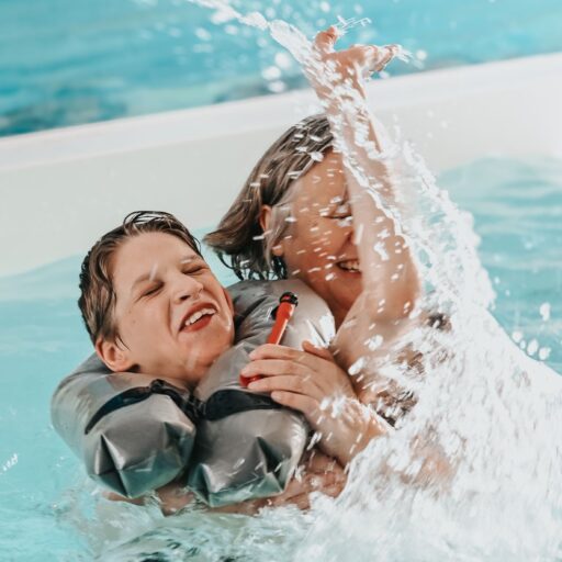 Eine ältere Frau planscht mit ihrem Enkel in einem Schwimmbad, der Wasser spritzt, beide lachen.